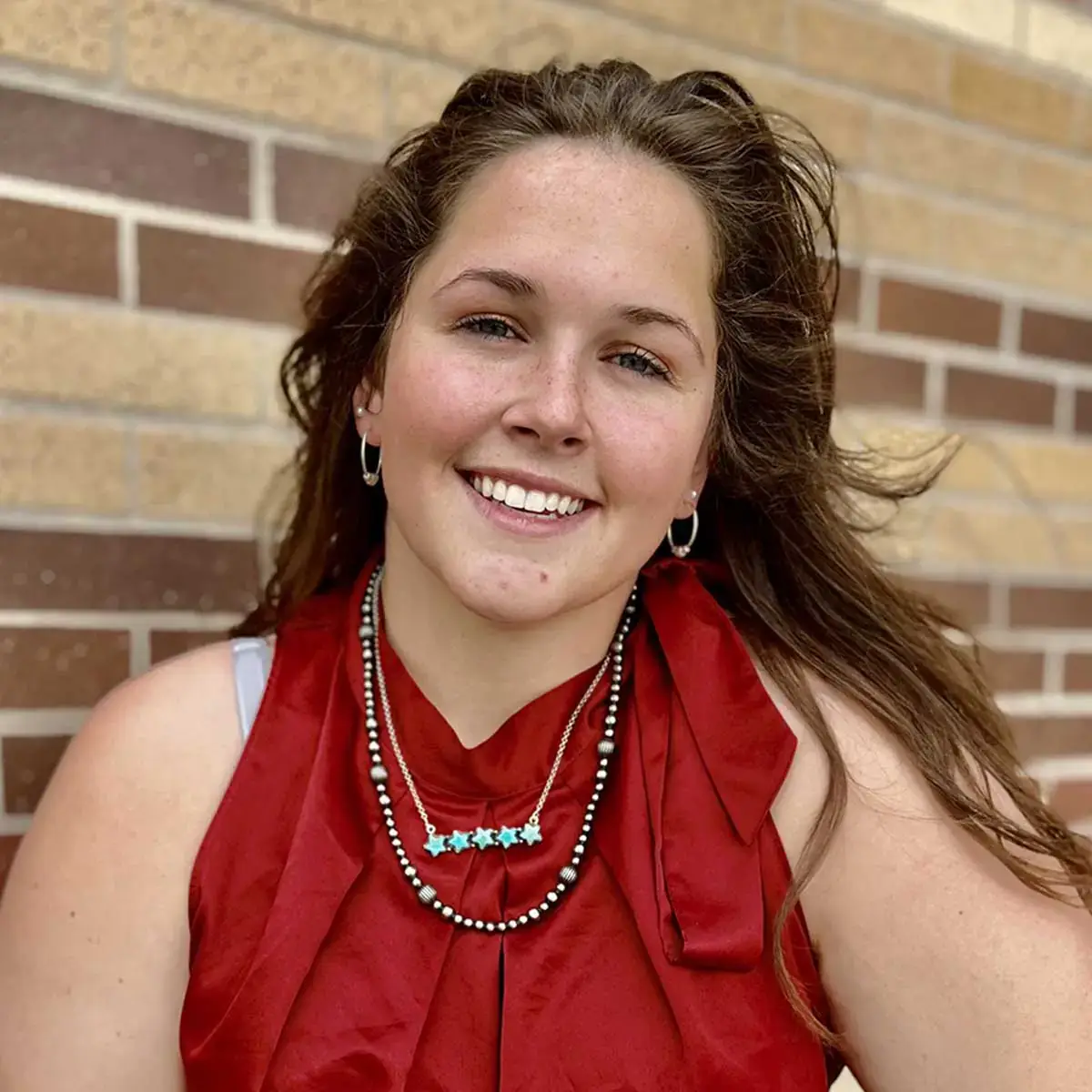 A person wearing a red top and jewelry smiling outdoors with a brick wall in the background.