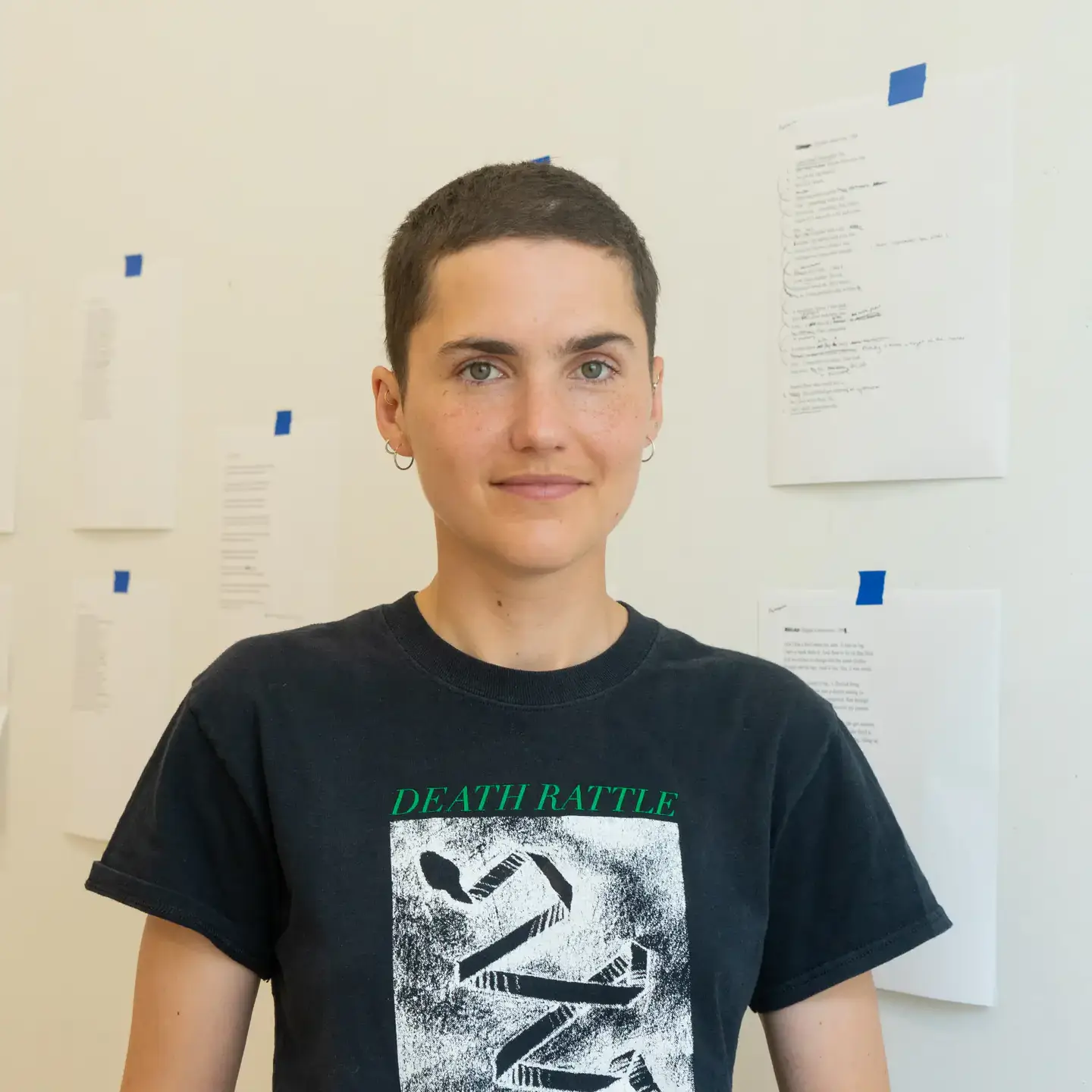 A person stands in a well-lit room with art pieces and text sheets taped on the wall behind them.