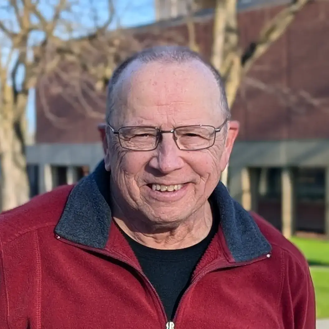 A person wearing a red zippered sweater smiling warmly, standing outdoors with trees and buildings in the background.
