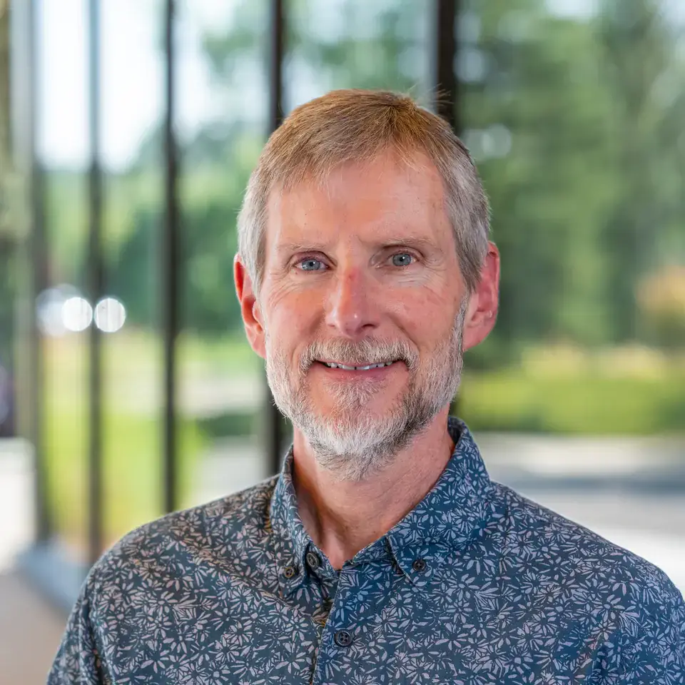 A person with light hair and beard is smiling in front of a window overlooking greenery, wearing a patterned shirt.
