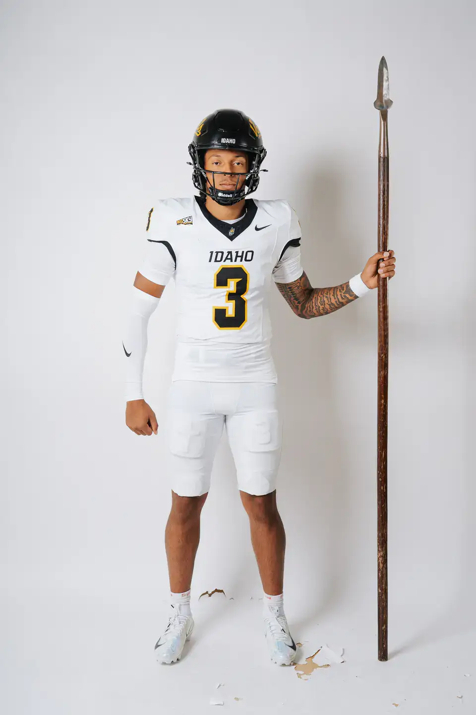 Football player wearing white unform holding a spear in his left hand standing in front of a white background.