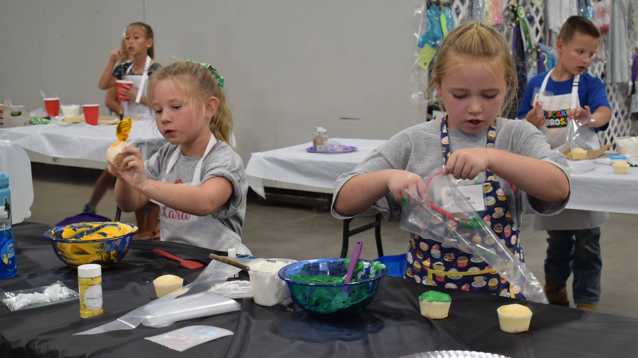 Two girls decorate cupcakes with orange and green icing, working on a long table with a black table cloth, while a boy and a girl work on their cupcakes at tables behind them.