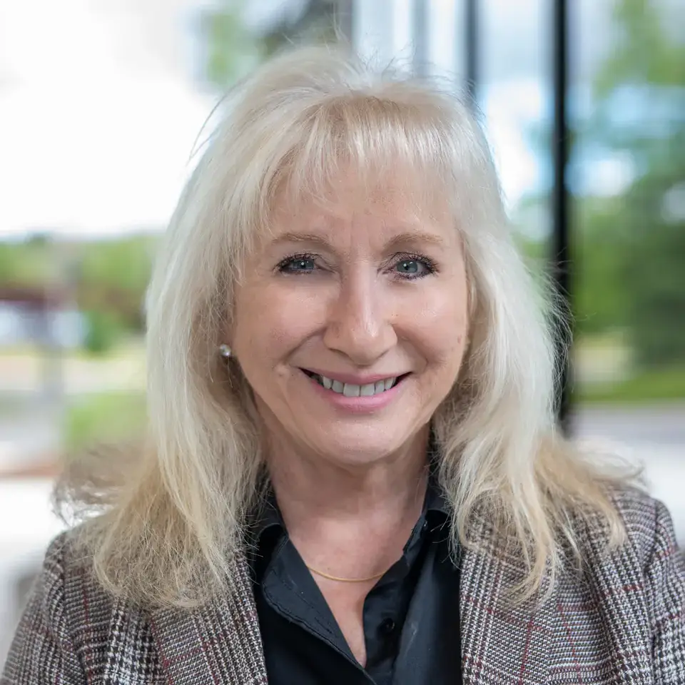 A smiling individual with long blonde hair wearing a plaid blazer and black shirt, seated with a blurred outdoor background.