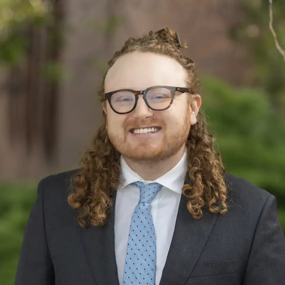 Person with long curly red hair, glasses, dressed in a suit and tie, outdoors with greenery in the background.