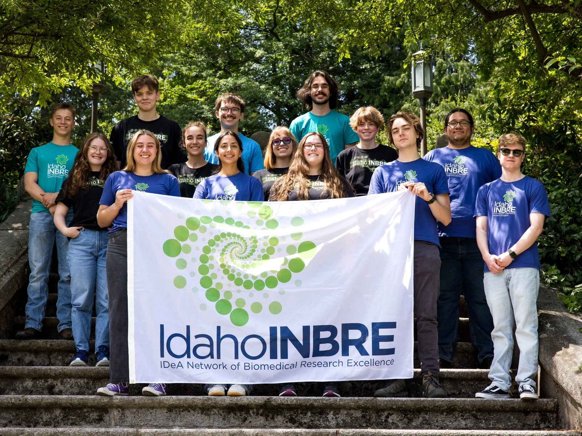 A group of 14 stands on stairs surrounded by green foliage holding a sign that says Idaho INBRE