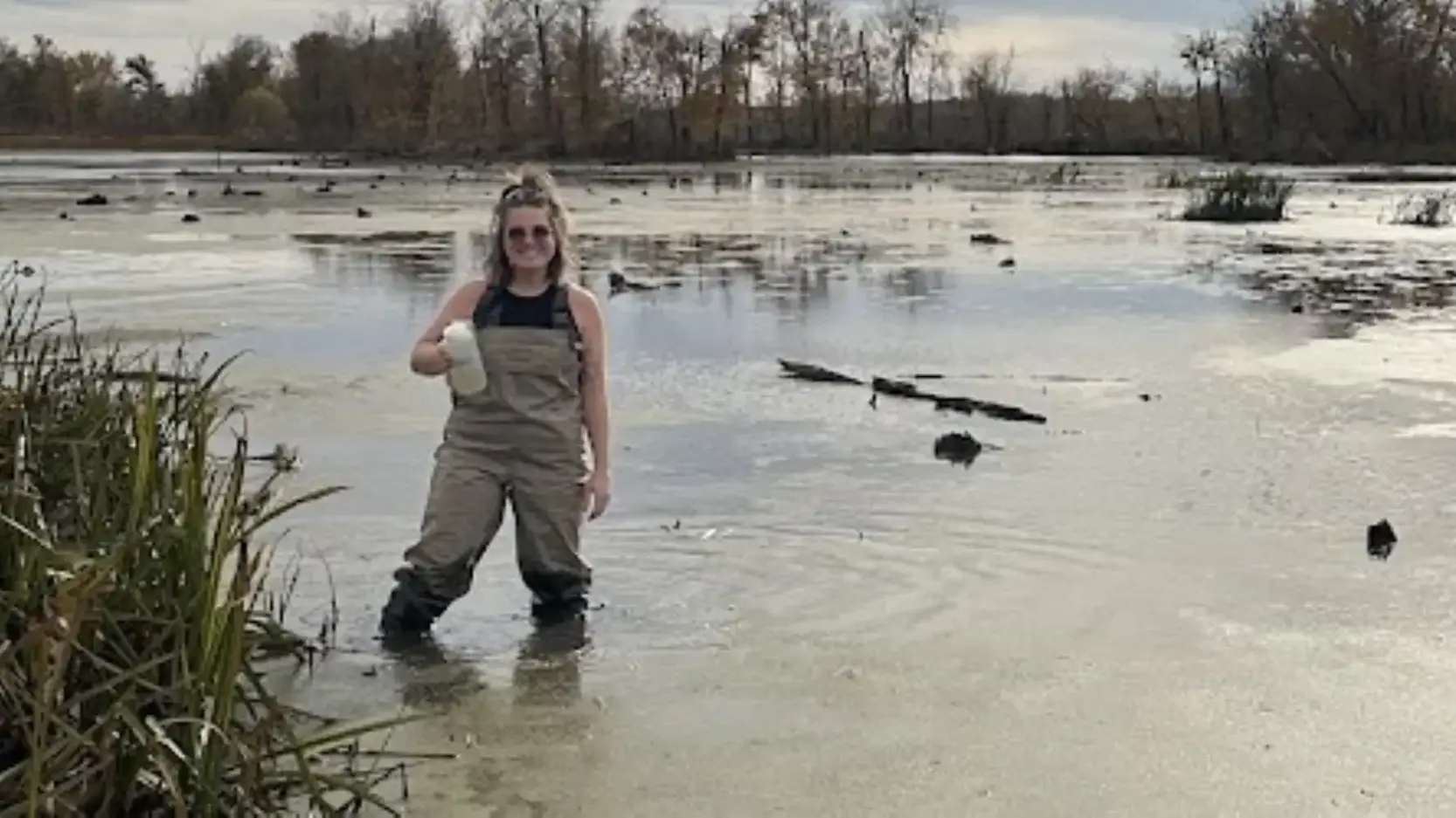 Woman wearing waders stands in a pond holding a jar of water.