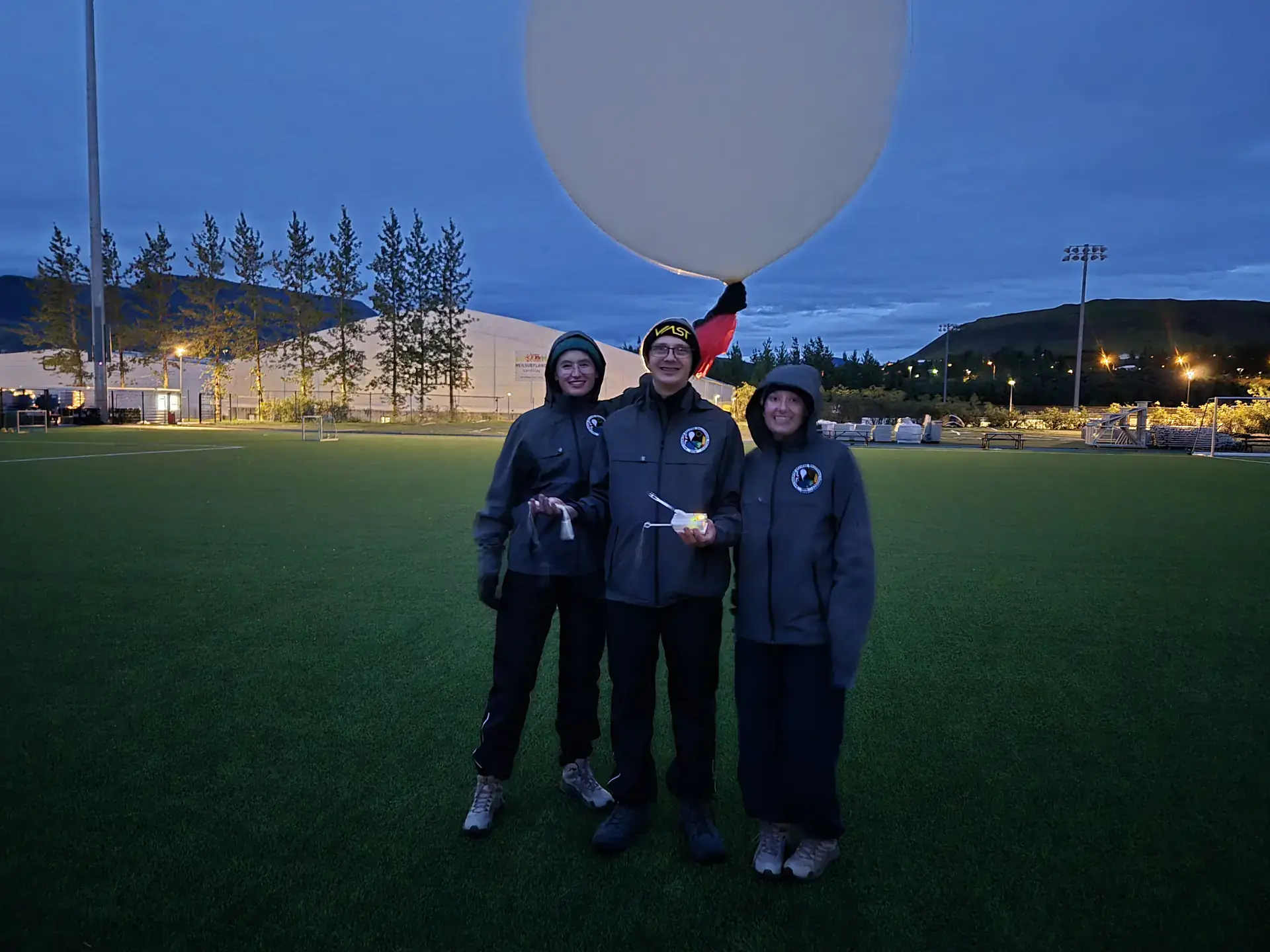 Three people in jackets stand on a field at dusk beneath a large balloon, with mountains and lights behind them.