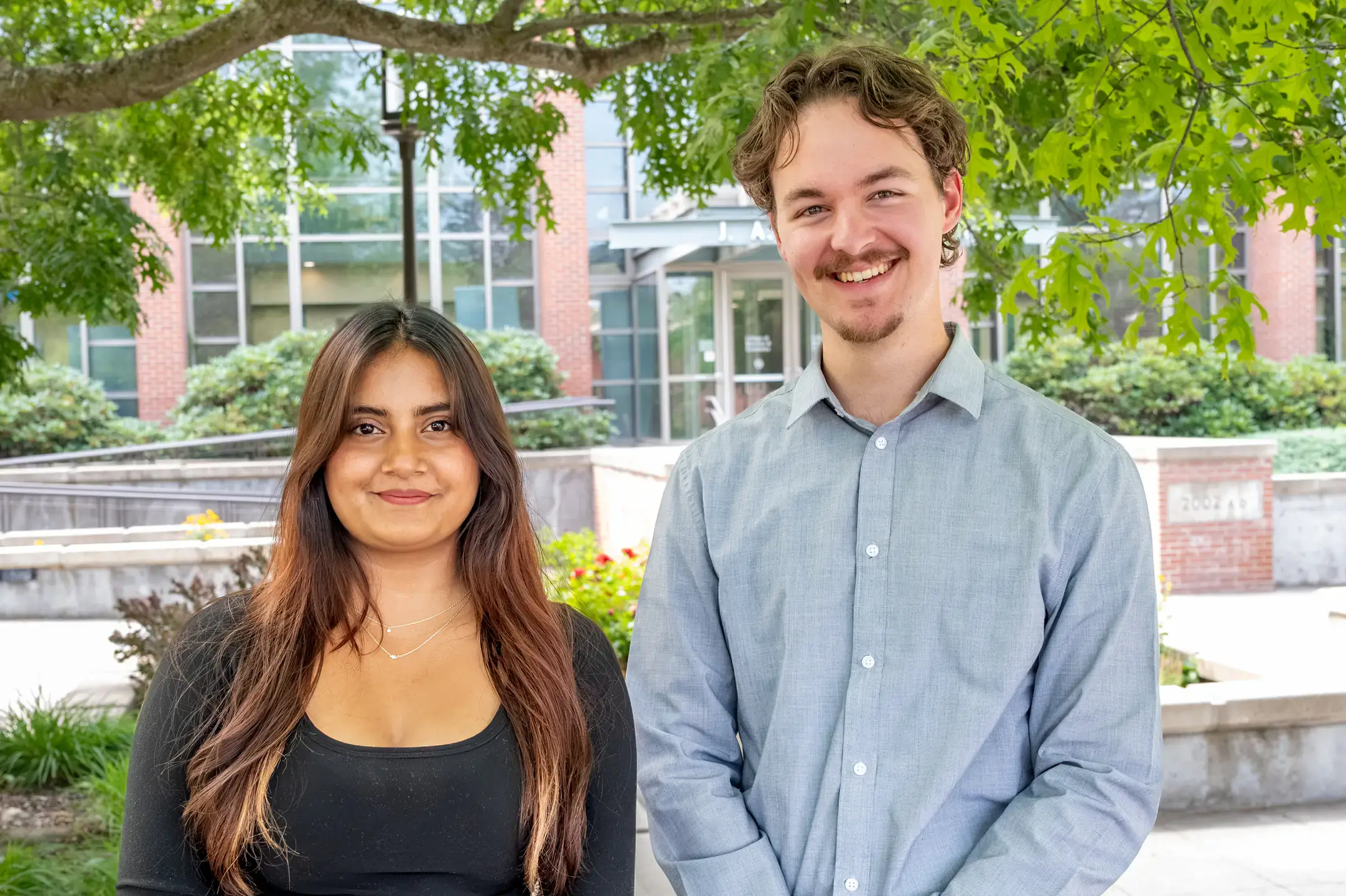 Two individuals standing outdoors in front of a building, smiling into the camera.