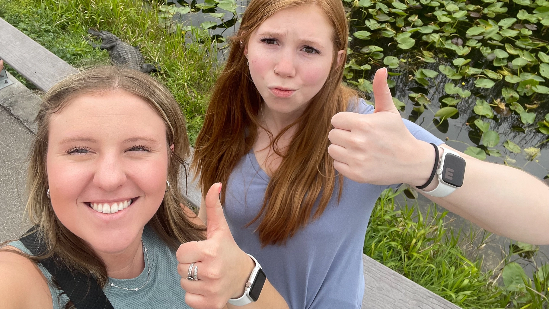 Two women give thumbs up in front of a swamp with an alligator in the background.