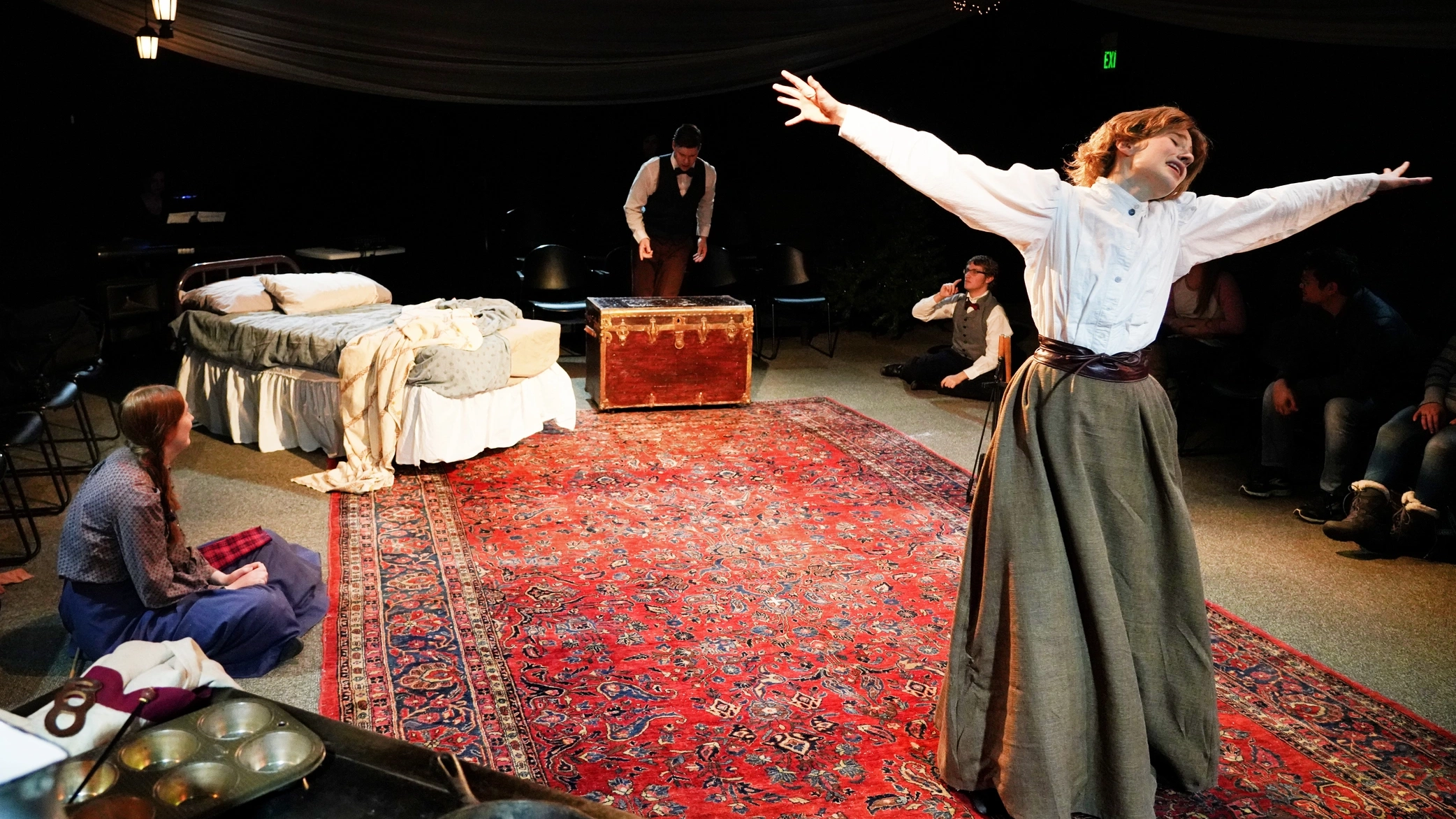 A woman standing with her arms out, with a bed and a man standing on a chest in the background