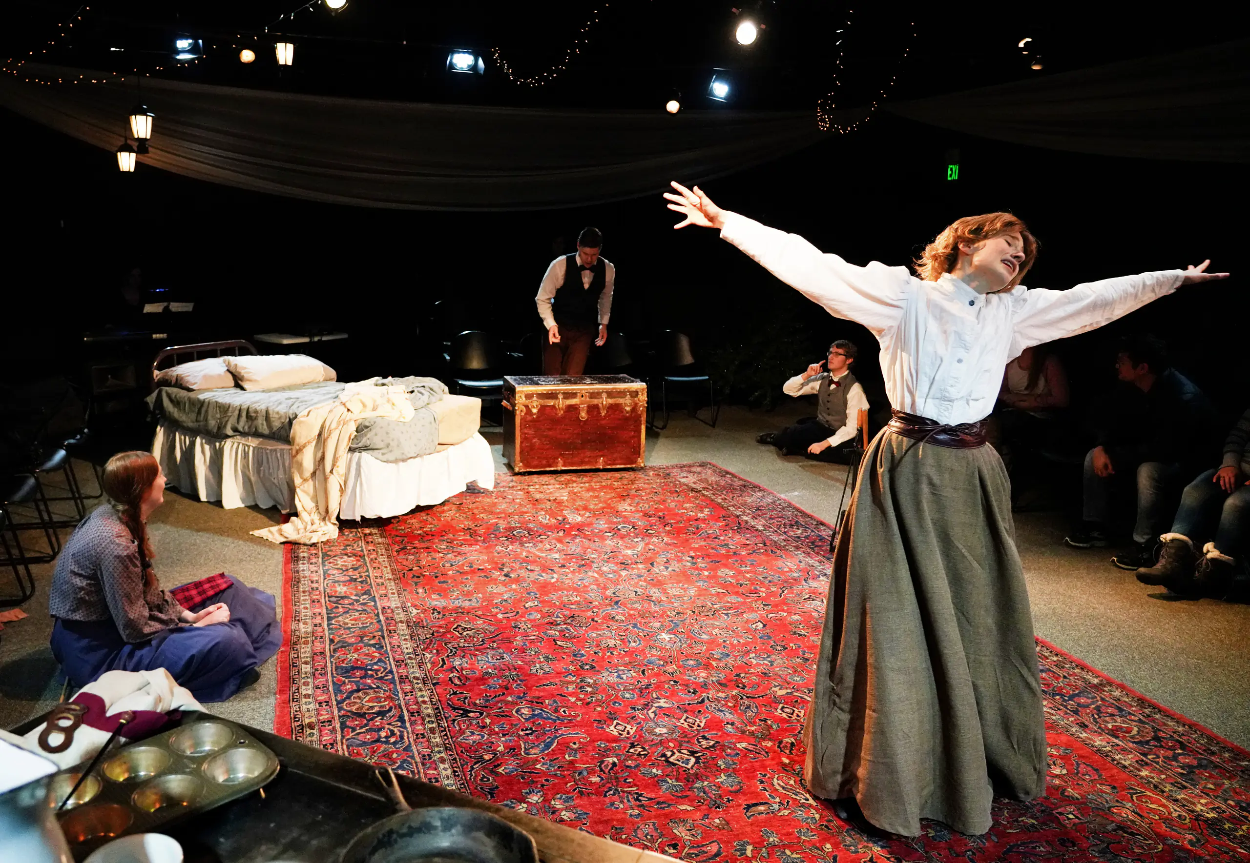 A woman standing with her arms out, with a bed and a man standing on a chest in the background