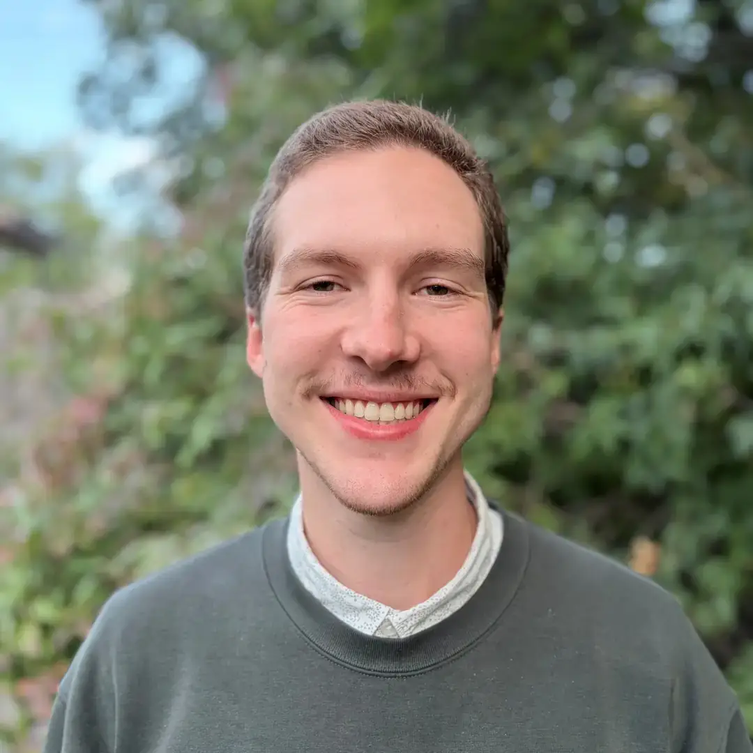 Smiling young man in a gray sweatshirt outdoors, with a blurred green background.