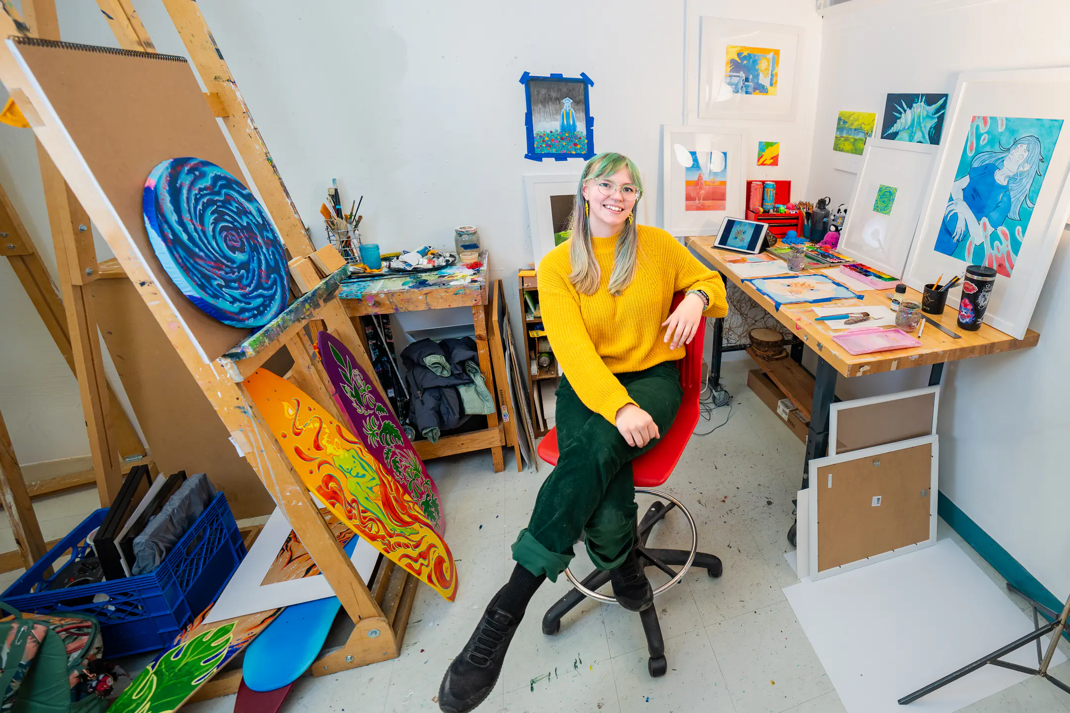 Kieran Heywood wearing a yellow top, sitting in chair amongst projects in their art studio.