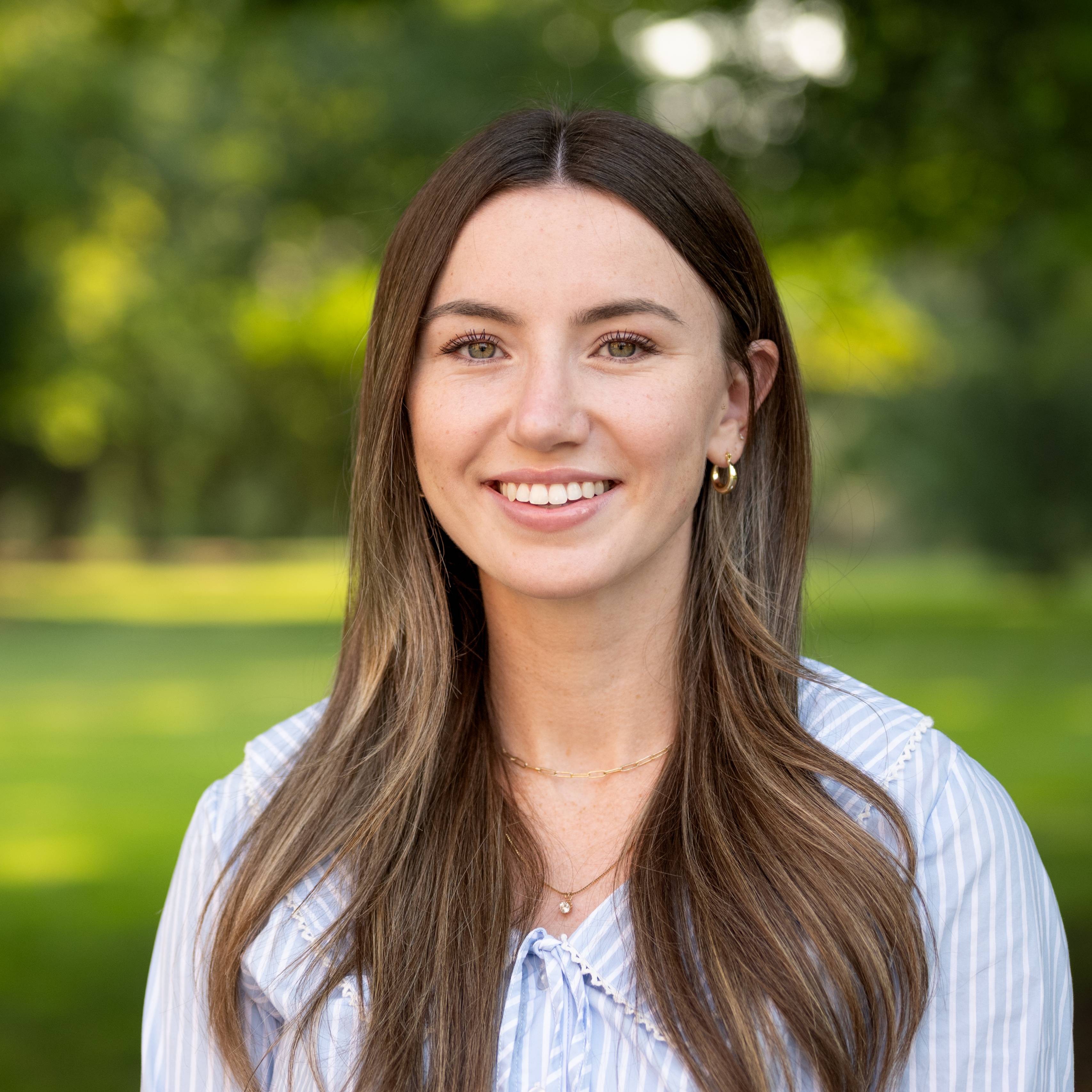 Smiling woman with long brown hair in a light blouse standing outdoors in a park-like setting with green trees and grass.