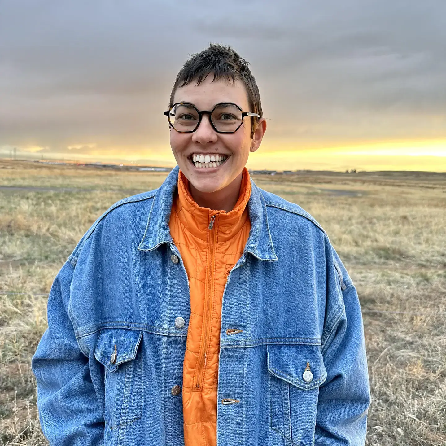 Person smiling outdoors in a grassy field during sunset, wearing glasses and a denim jacket over an orange shirt.