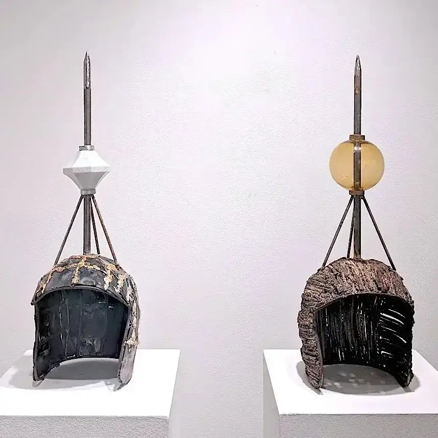 Two weathered helmet sculptures on white pedestals, each topped with a tall spike and geometric ornament.