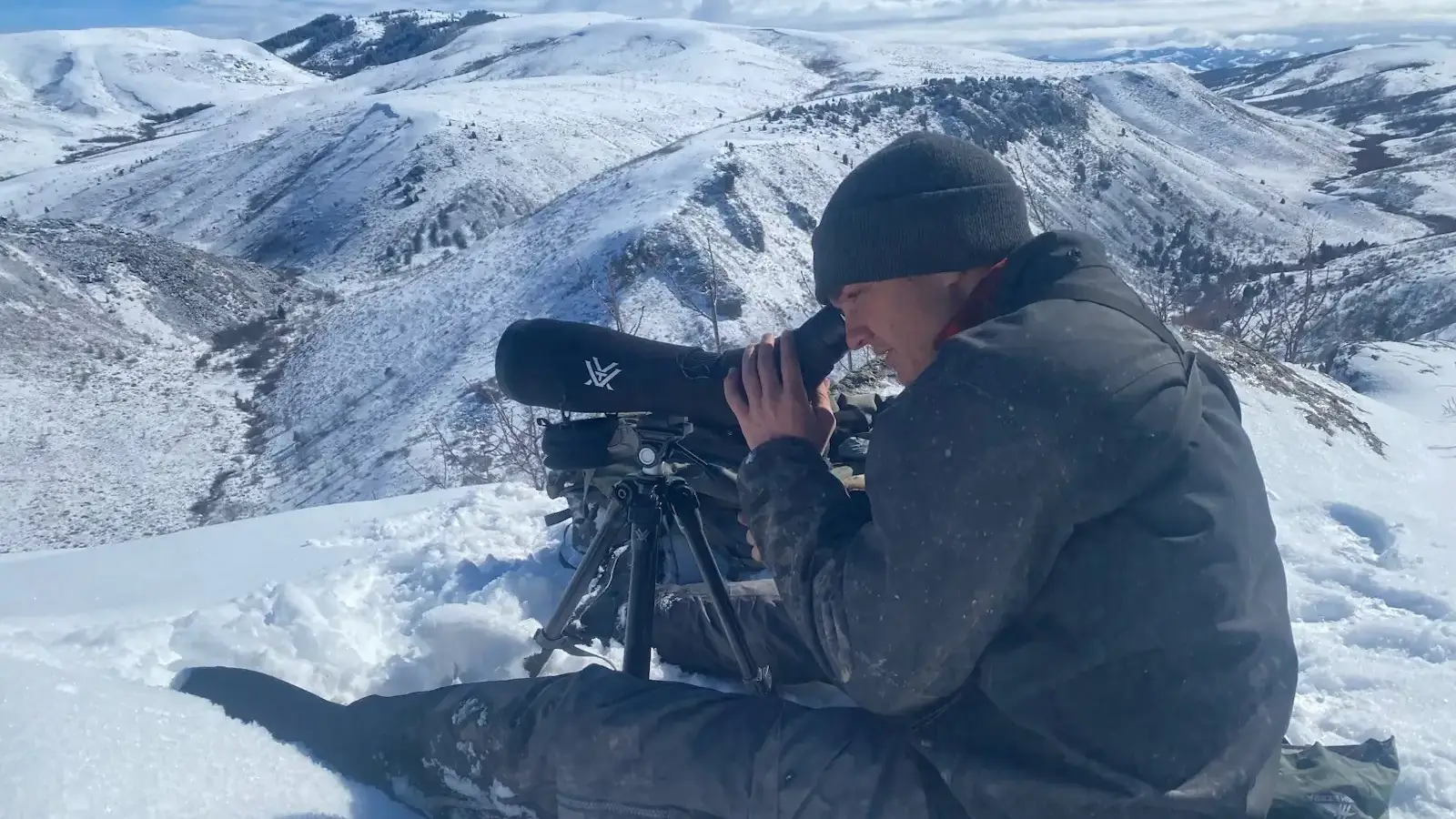 A man in an arctic setting looks through a telescope device to observe something in the distance.  
