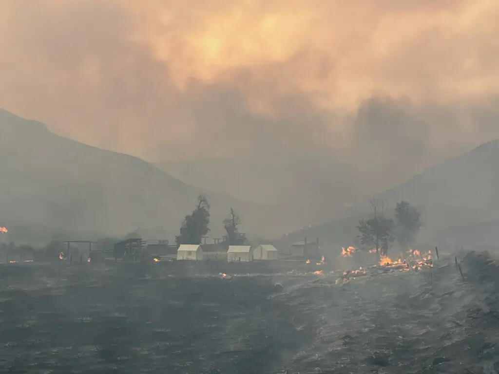 A landscape photograph of a heavily smokey area of rangeland with white wall tents and burning trees in the background.