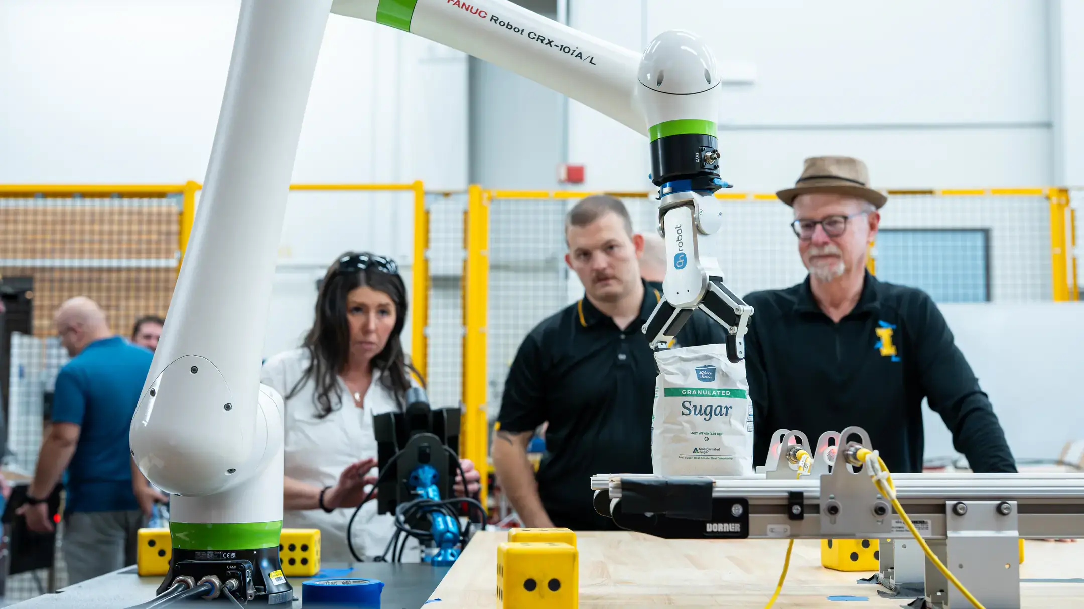 Two students and a professor in a computer lab, controlling a robotic arm to lift bags of sugar like a crane.