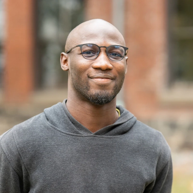 A man smiles outdoors with a building in the background.