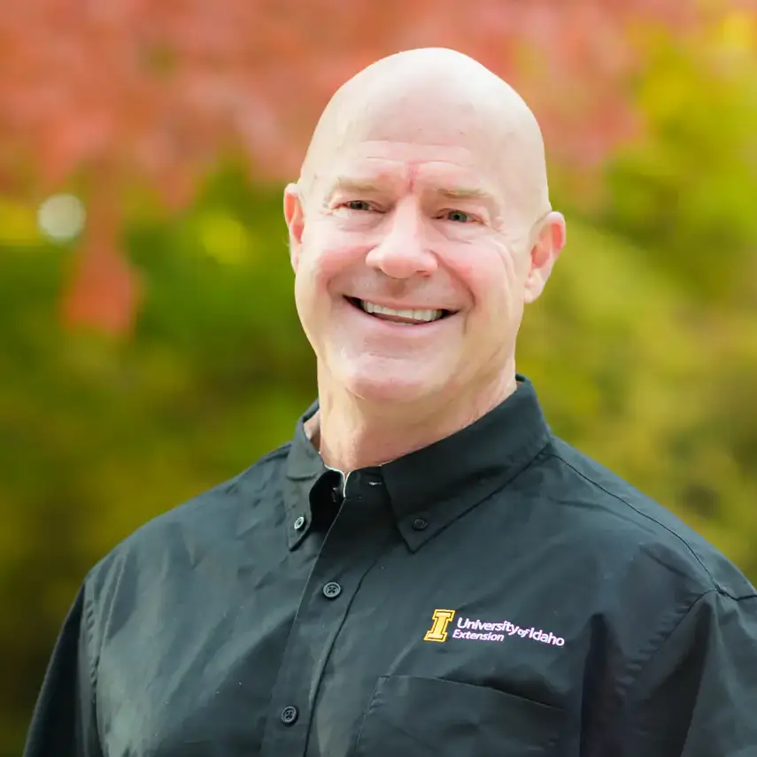 Smiling man in a University of Idaho Extension shirt standing outdoors before colorful autumn trees.