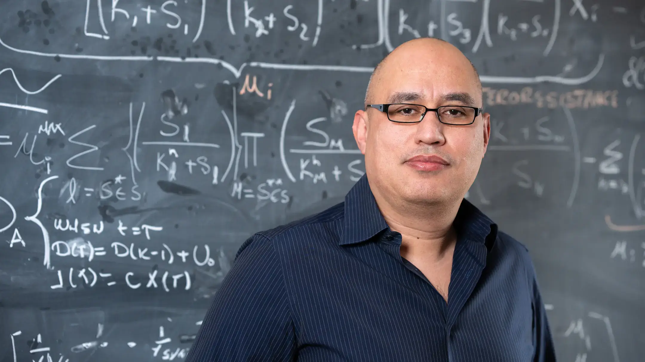 Portrait of Assistant Professor of Mathematics Esteban Hernandez-Vargas in front of a chalkboard with math equations models to study the mitigation of viral infections.