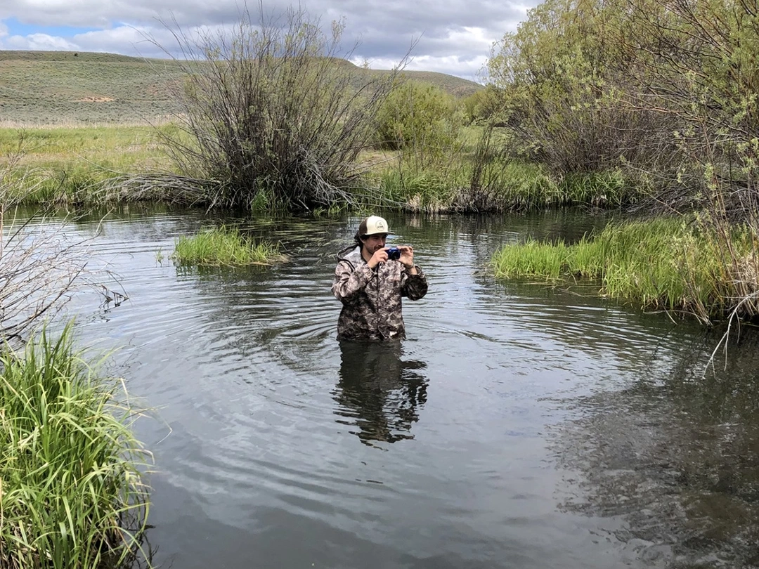 A man stands in the middle of a rangeland pond holding a camera.