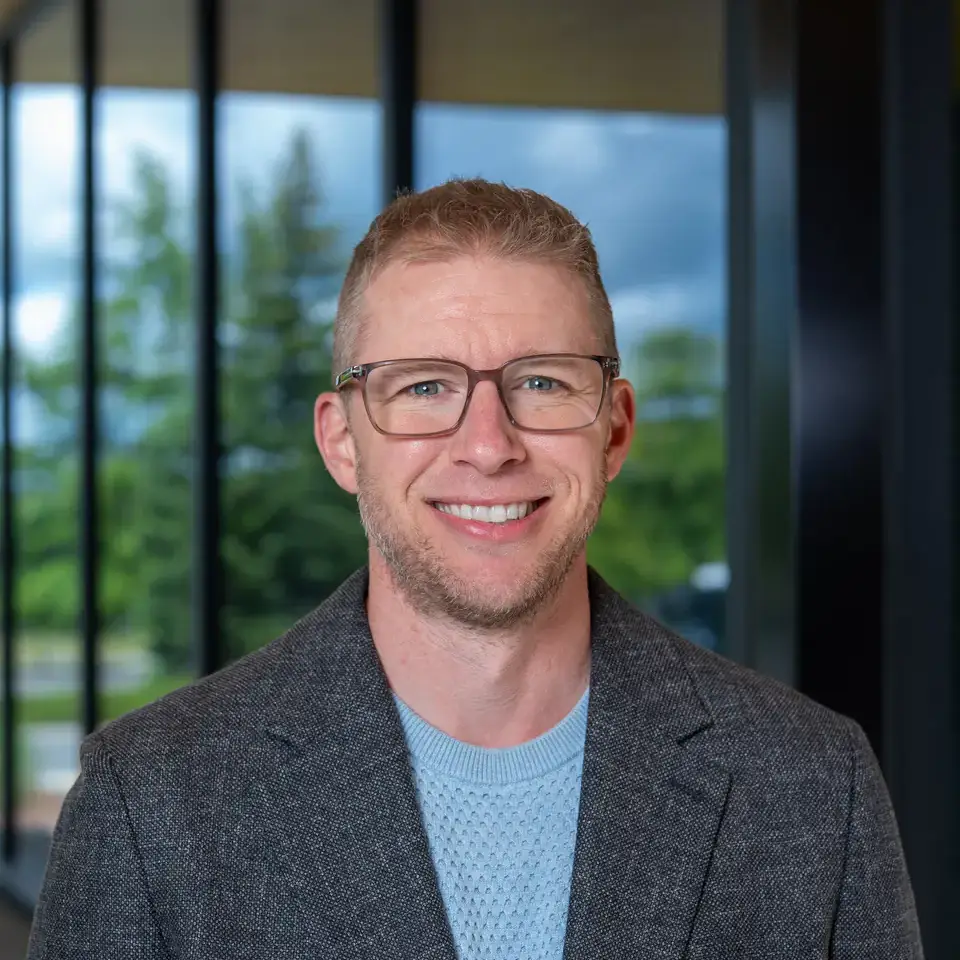 A man wearing glasses and a gray suit jacket over a light blue sweater, smiling and standing indoors near a window.