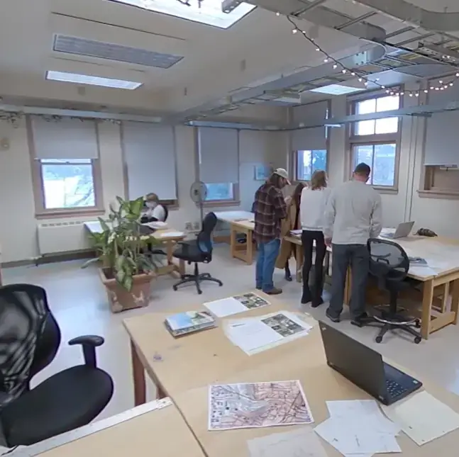 People collaborate in a bright studio with desks, maps, laptops, and papers spread across the tables.