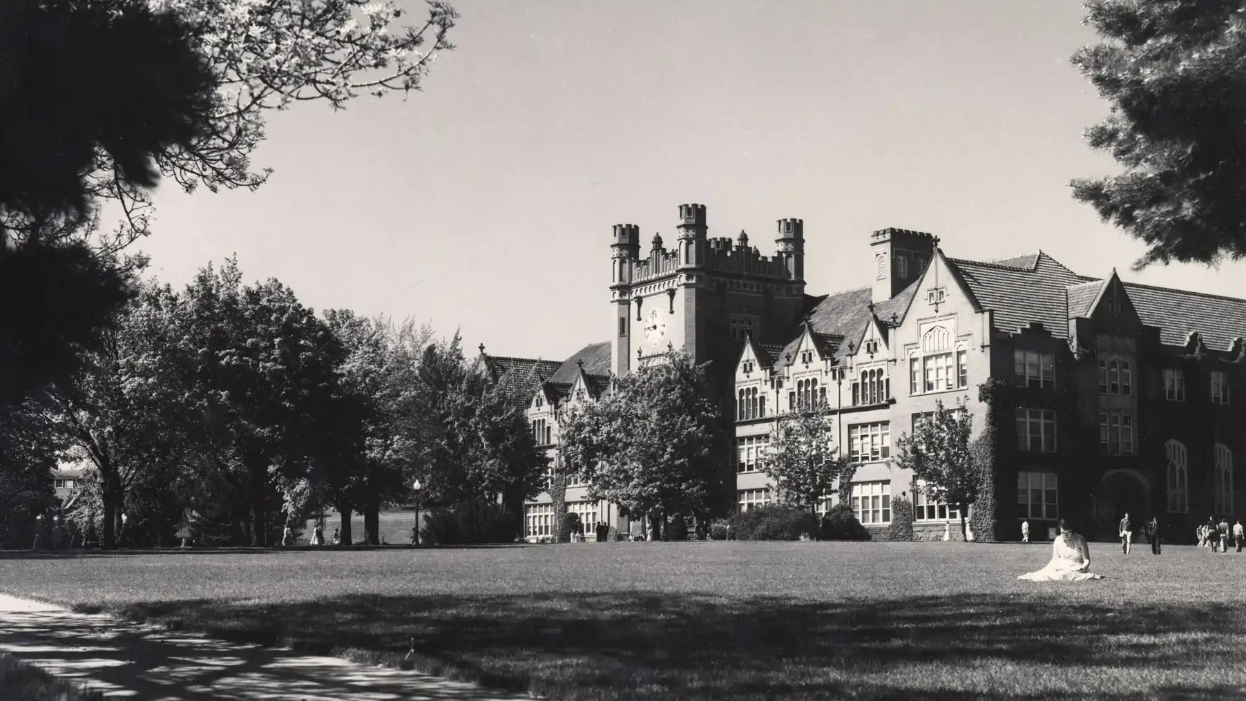Black-and-white photo of a large Gothic-style campus building across a broad lawn, framed by trees.