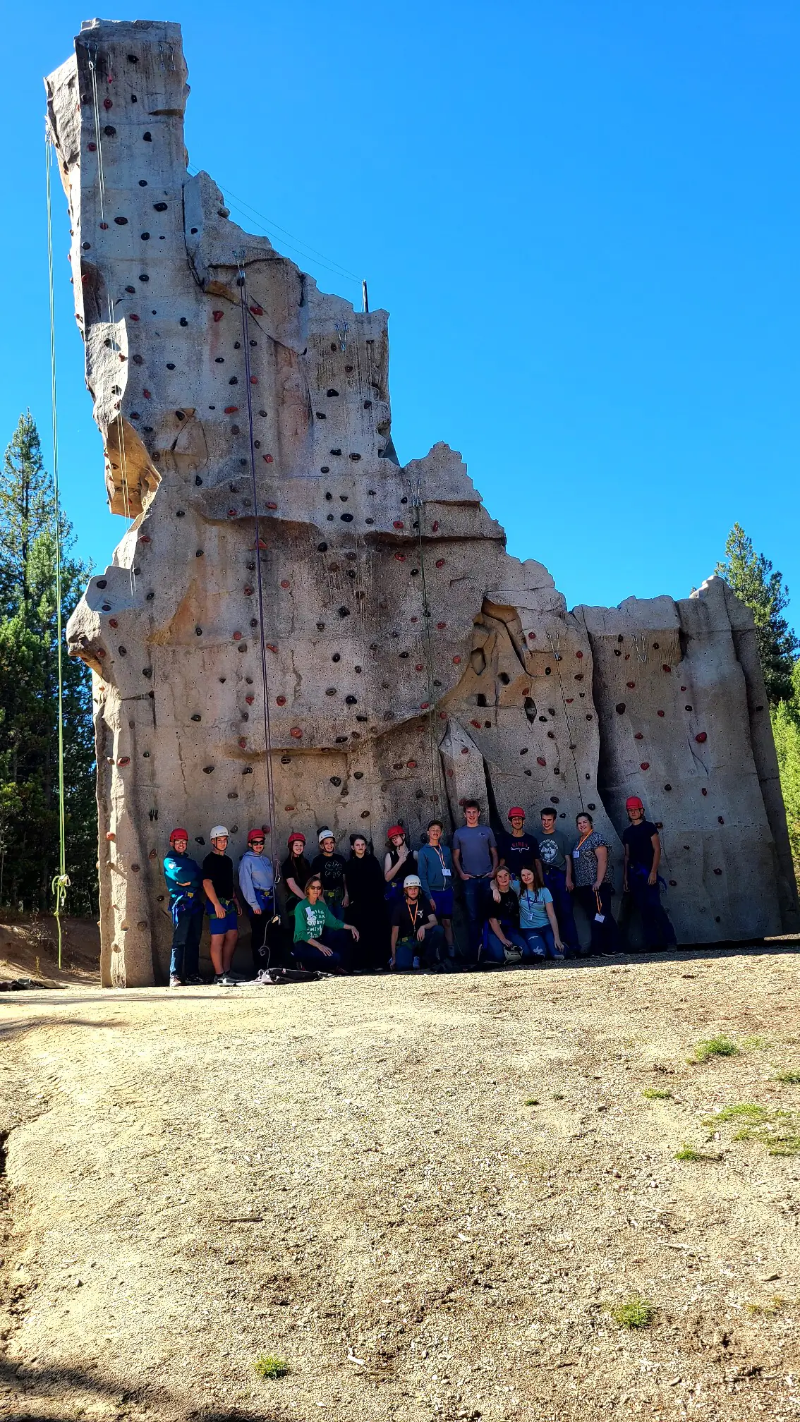  A group of youth wearing rock climbing gear pose together at the base of a rock wall shaped like the state of Idaho.