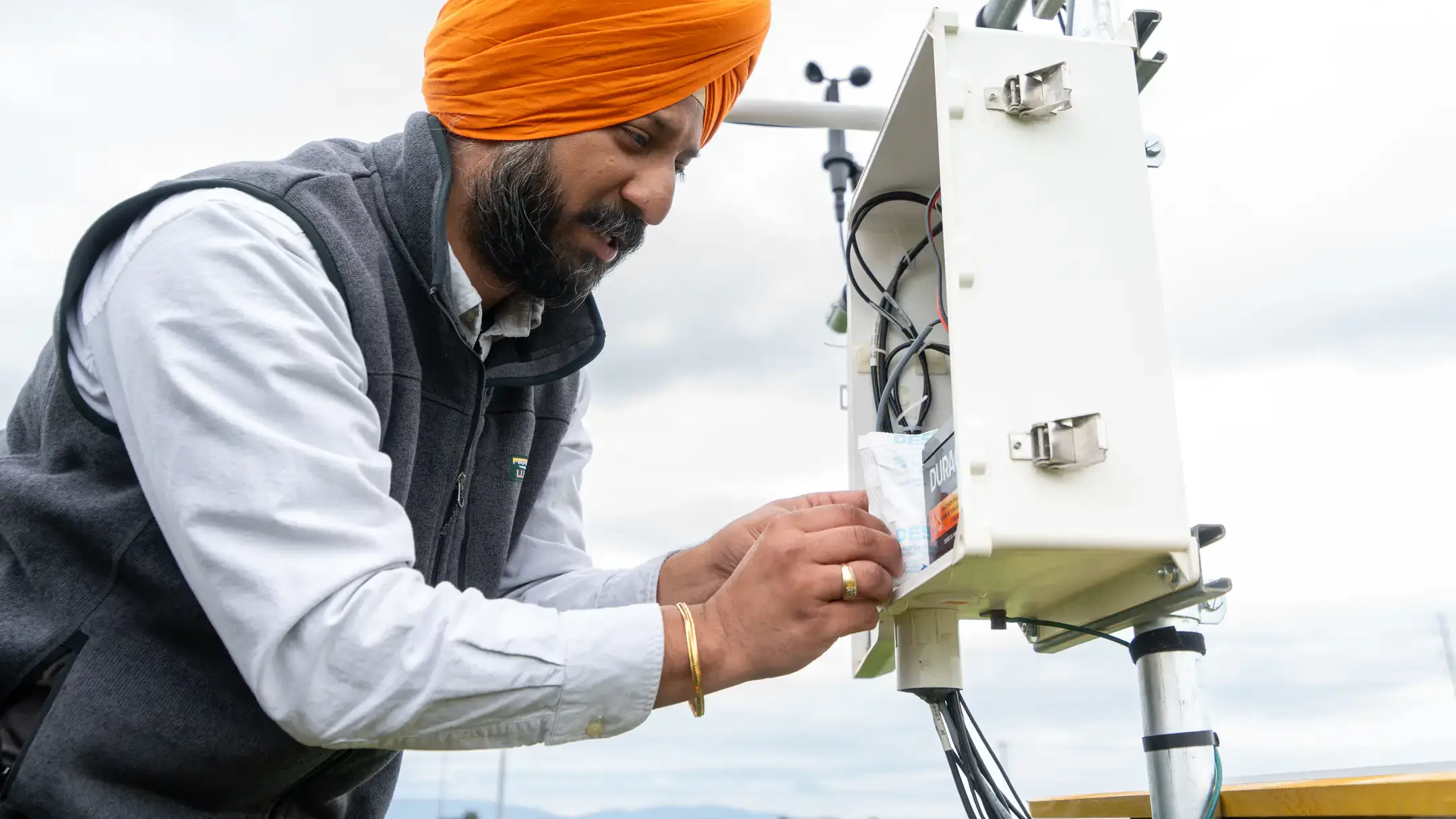 A professor works on a white, metal box mounted on a pole with wind-monitoring instruments.