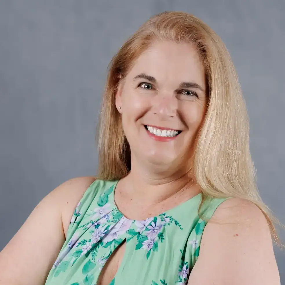 A smiling woman with long blonde hair wearing a green dress with a floral pattern. A neutral gray background.