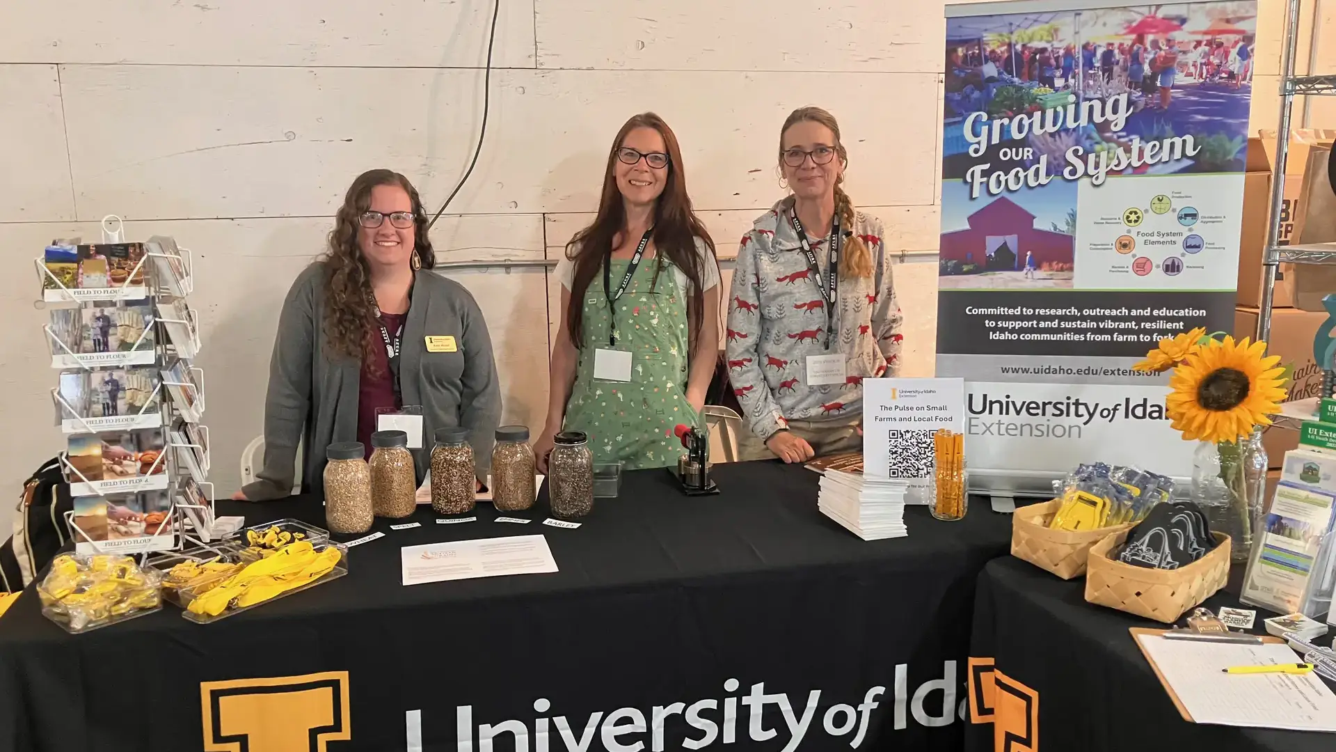 Three women stand behind a University of Idaho Extension booth with agricultural displays and brochures.