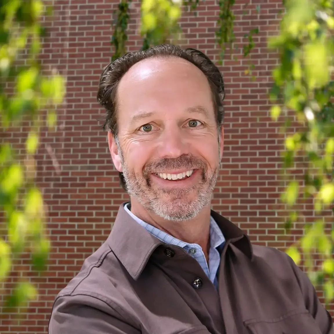 Richard Briend, smiling in front of a brick wall under trees.