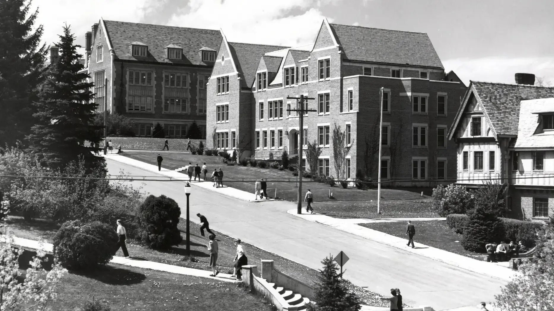 Black-and-white photo of students walking between large campus buildings along a curved street.