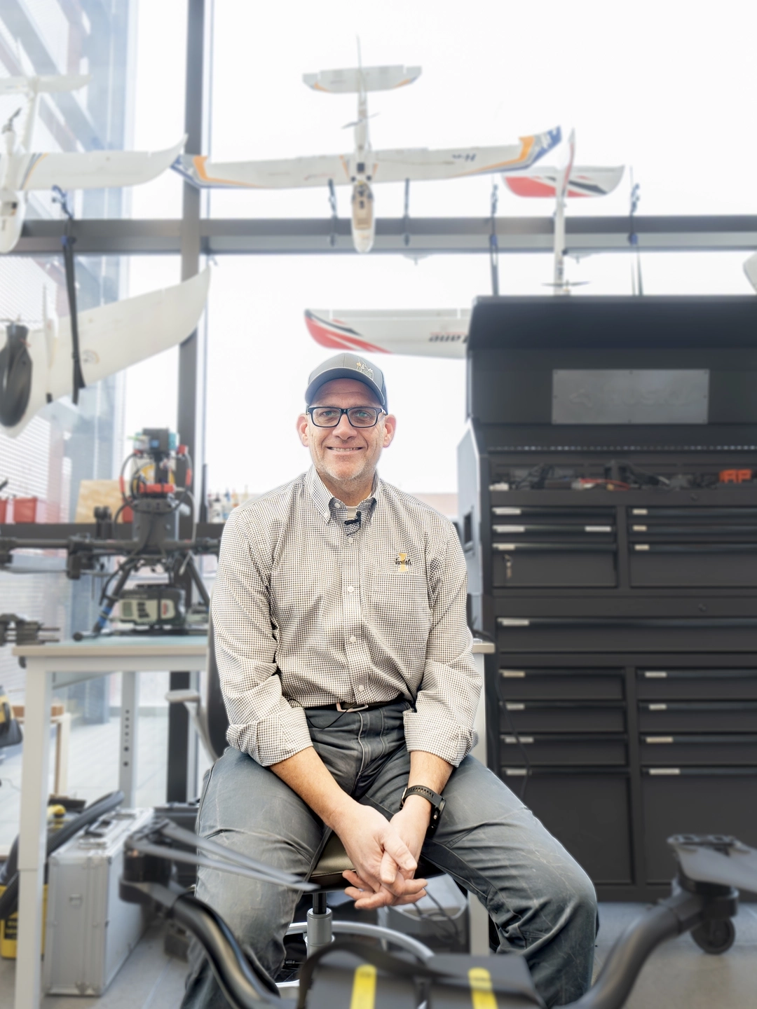 A man sites on a stool under a series of drones hanging from the ceiling.