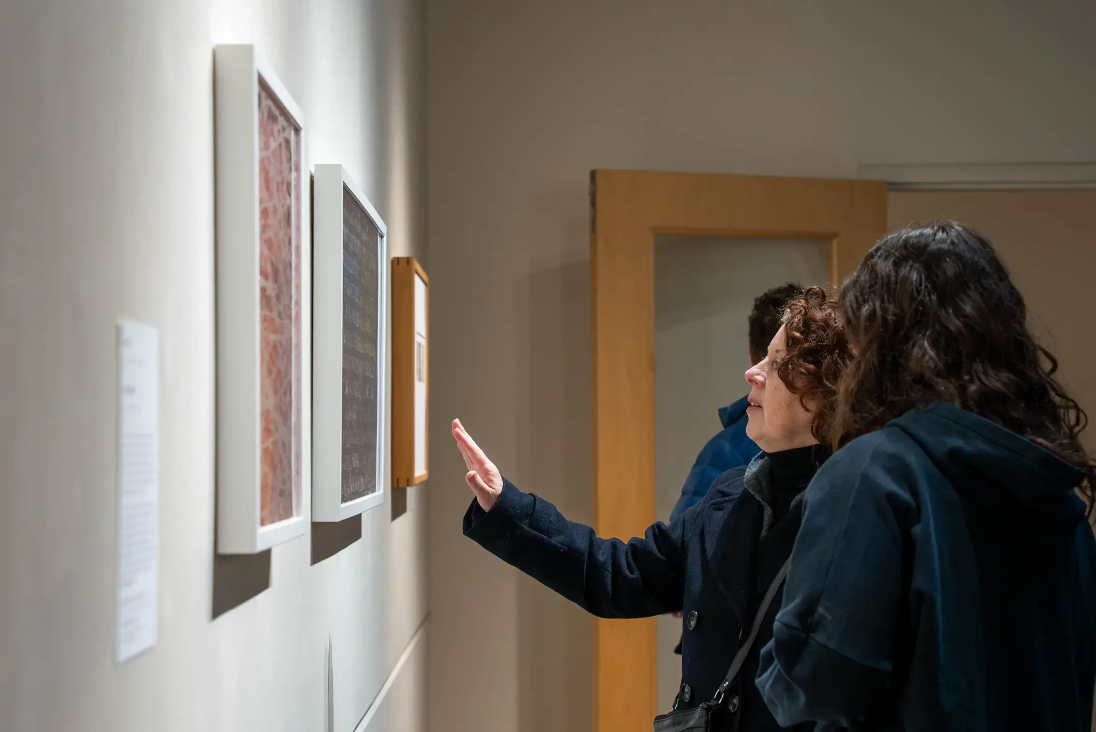 Two individuals viewing framed artworks displayed on a brightly lit gallery wall.