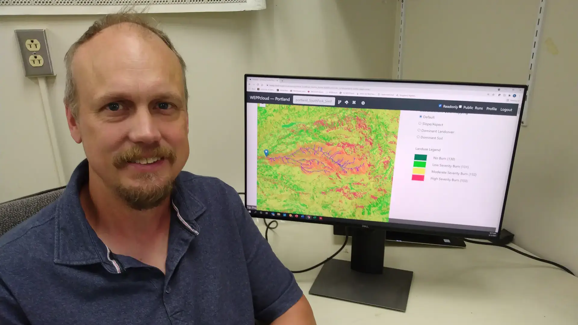 A man poses by a computer with a color-coded map on the screen.