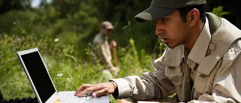 Man outdoors working on laptop taking notes in field