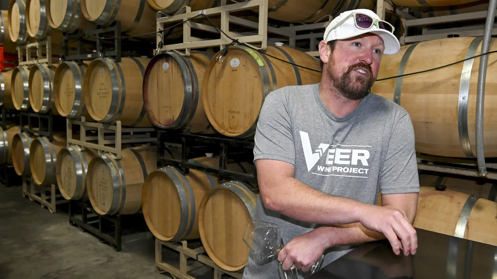 Person in a wine storage room with wooden barrels, wearing a cap and a shirt with "Veer Wine Project" written on it.