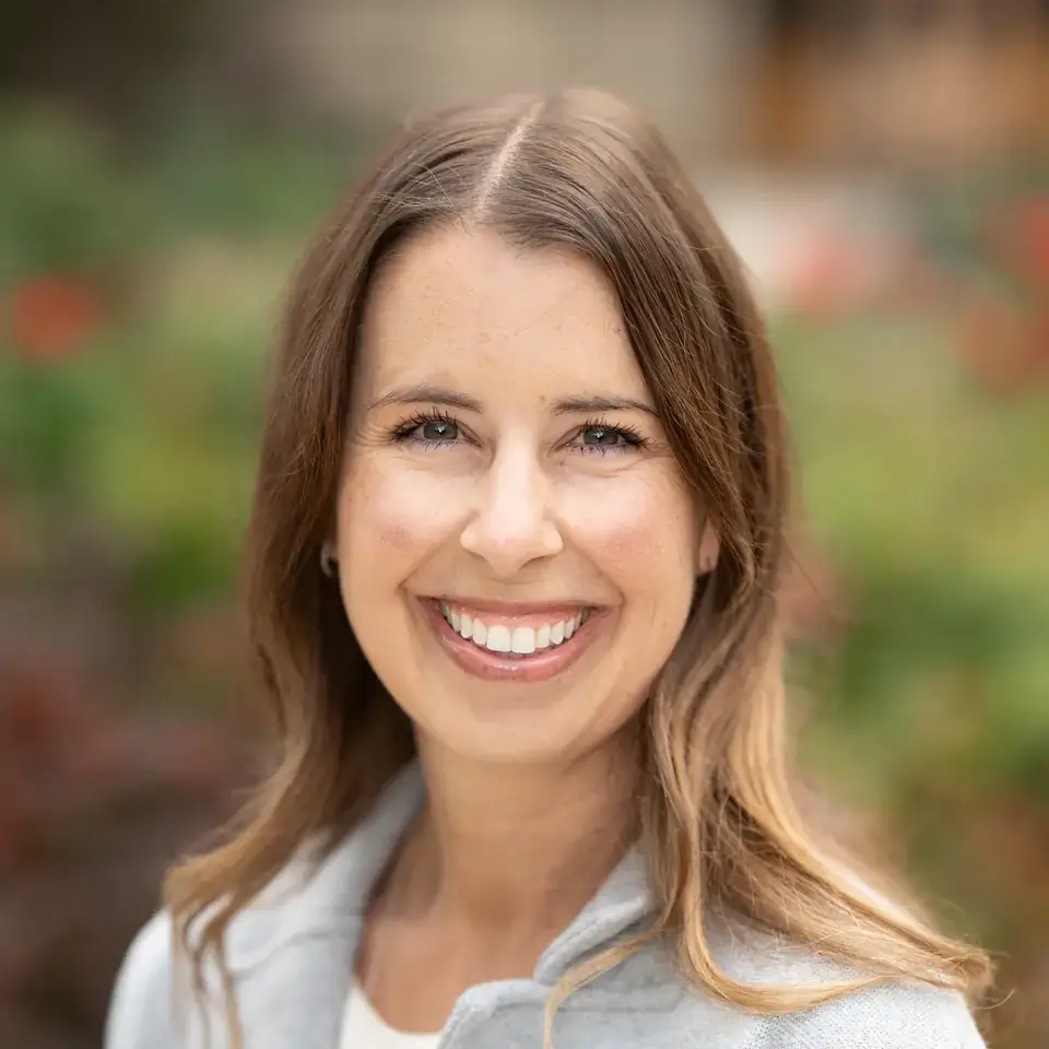 Smiling woman with long brown hair in a light gray jacket, photographed outdoors with a blurred garden background