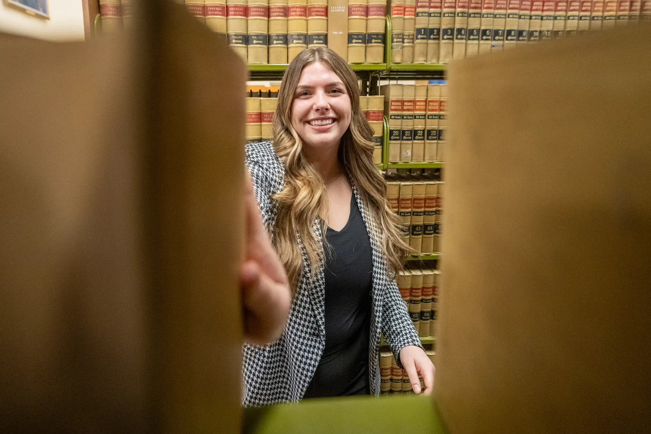 Professionally dressed smiling woman reaches for a book on a shelf.