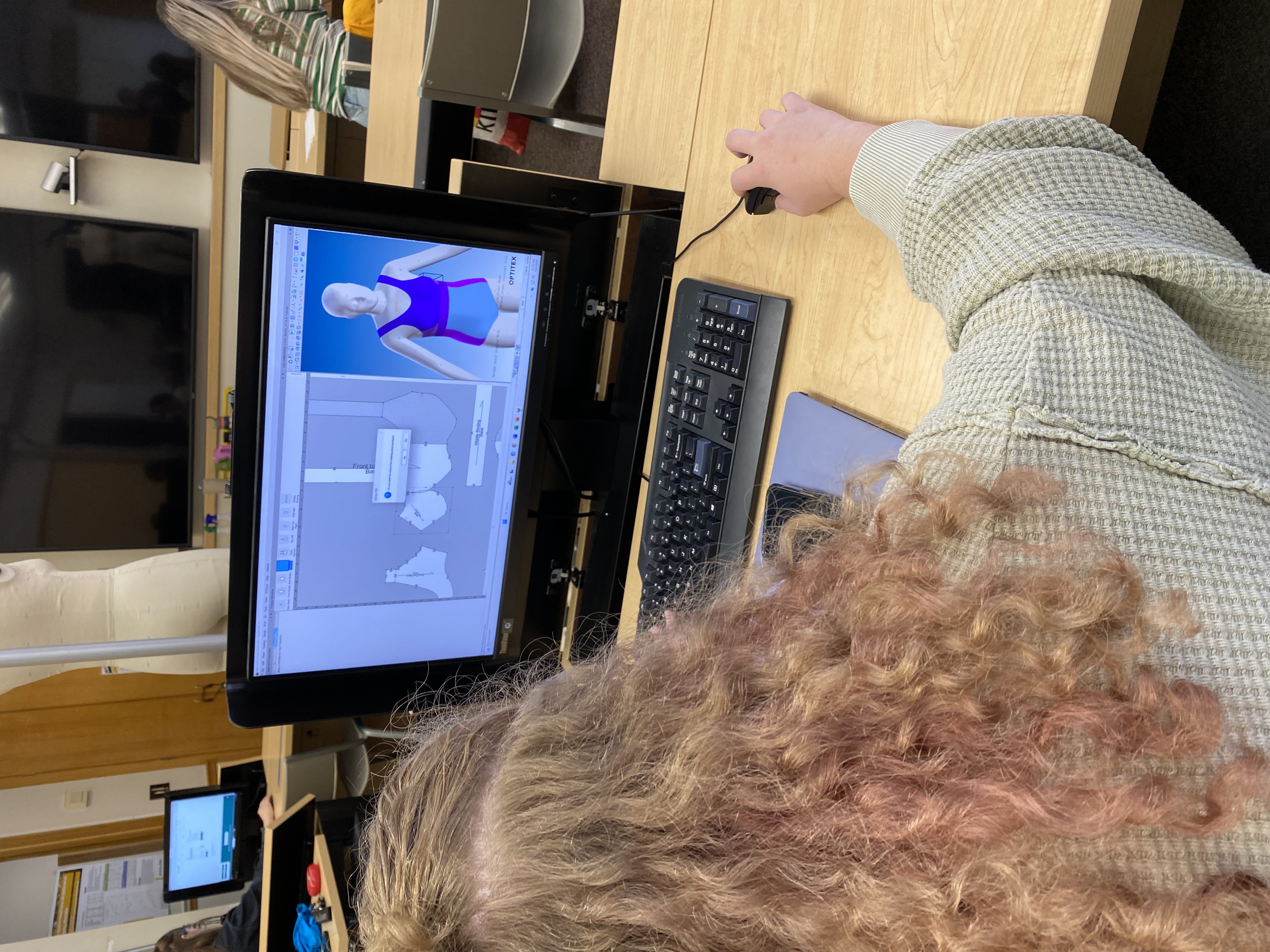 A person uses software for digital patternmaking on a computer screen, showing a garment pattern and a 3D model on display.