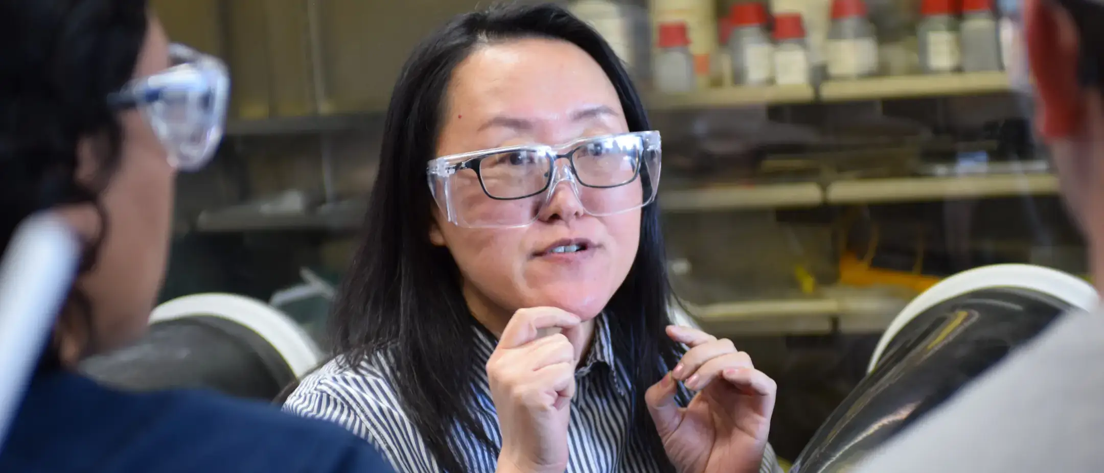 A person wearing safety goggles speaking to others in a laboratory setting, with shelves containing containers in the background.
