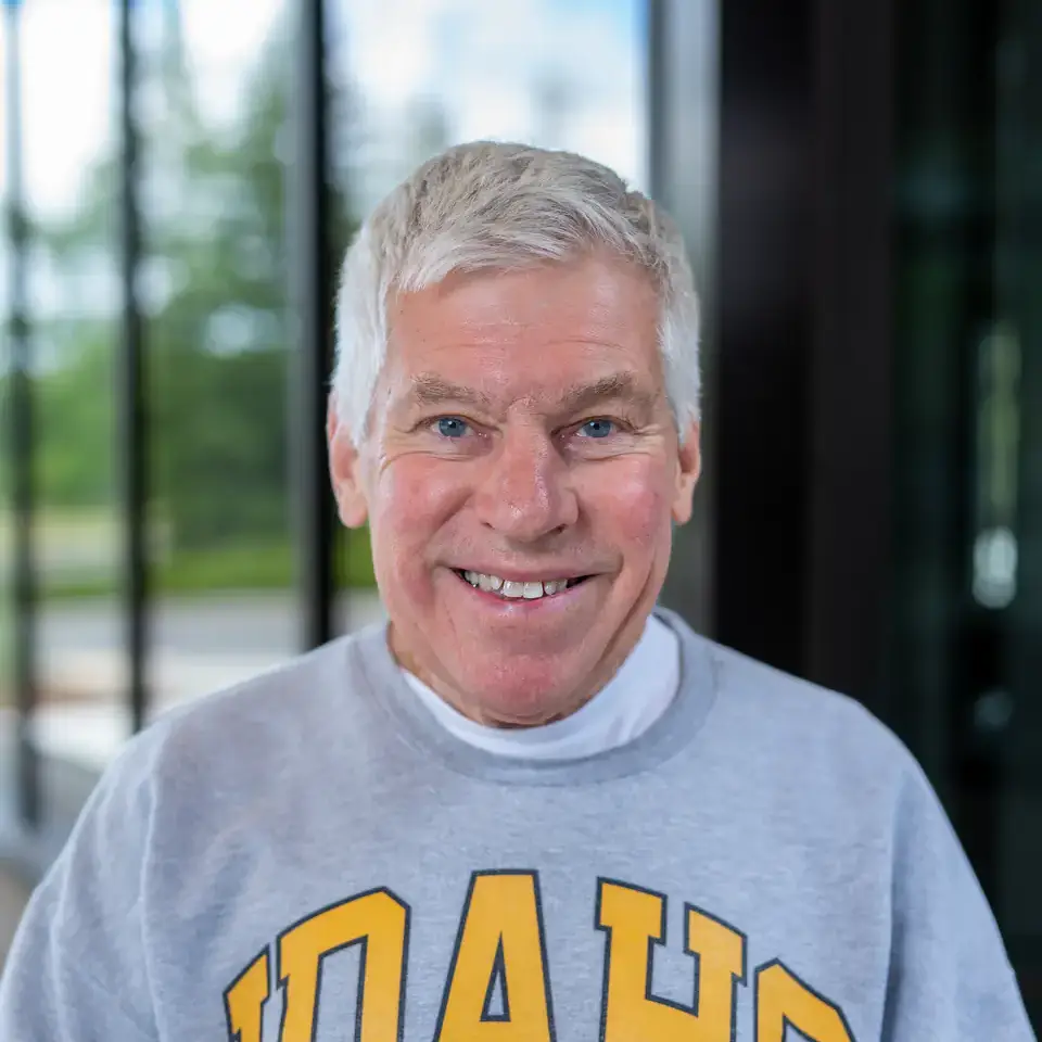 A person smiling, wearing a gray sweater with 'IDAHO' printed in yellow letters, standing in front of glass windows with trees visible outside.