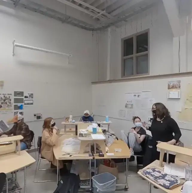 Students in a classroom working on projects and talking, with drawings and materials spread on desks.