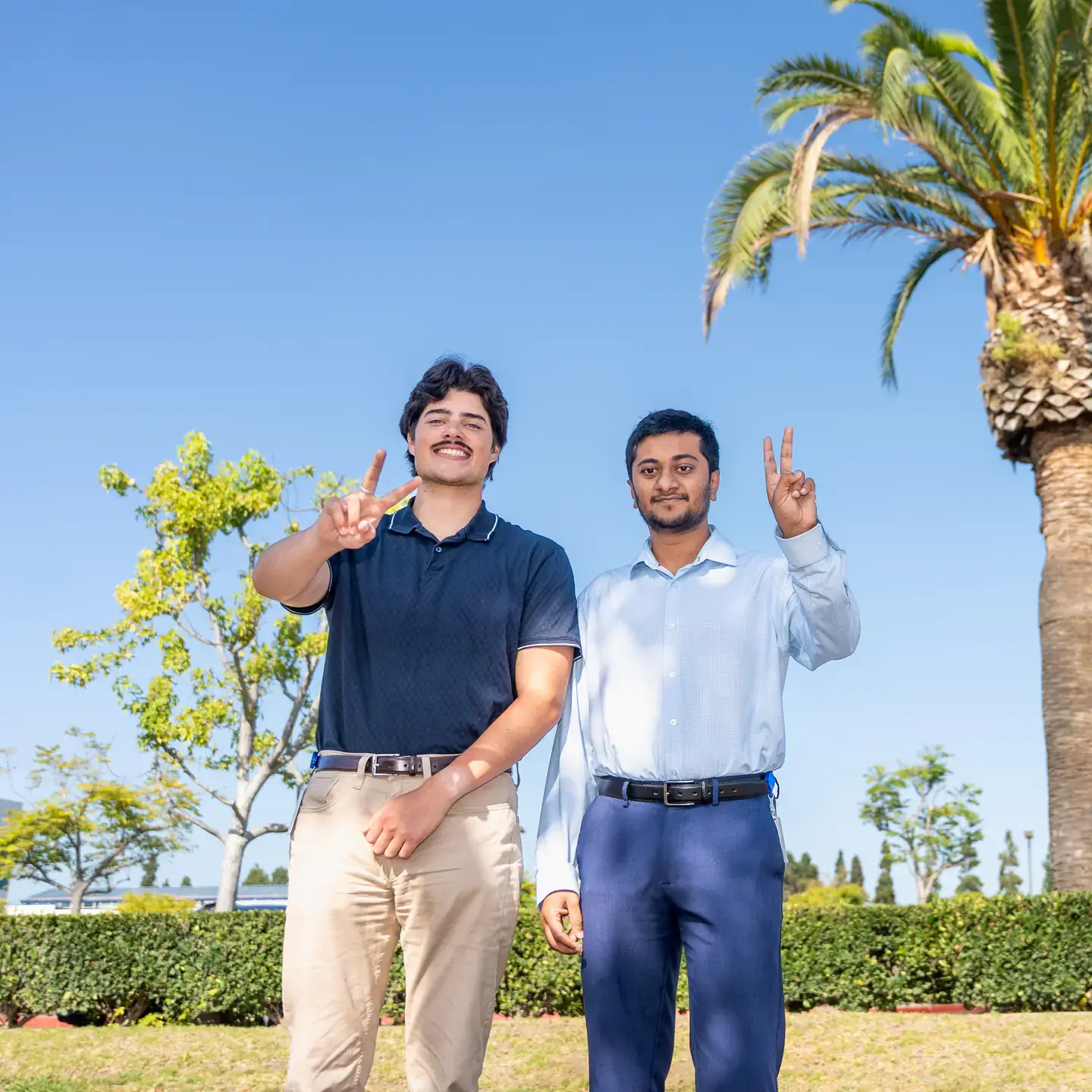 Two individuals stand outdoors under palm trees, smiling and gesturing towards the camera, with greenery and clear blue sky in the background.