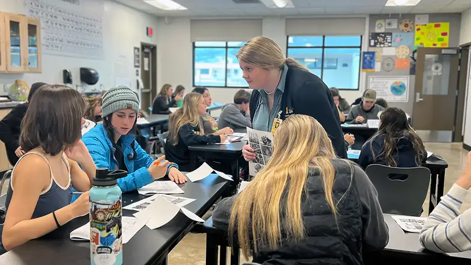 A female student teacher talks with a table full of high school students in a classroom.