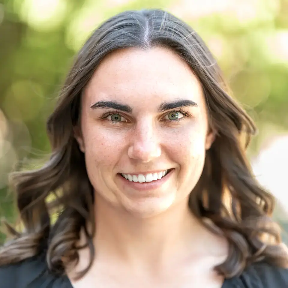 Smiling person with wavy hair posing outdoors with blurred greenery in the background, wearing a black top.